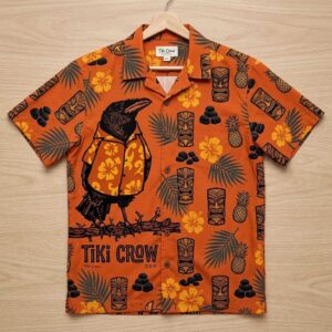 Aloha Crow wearing a Hawaiian t-shirt. Burnt orange tropical shirt with crow, tiki masks, pineapples, hibiscus flowers.